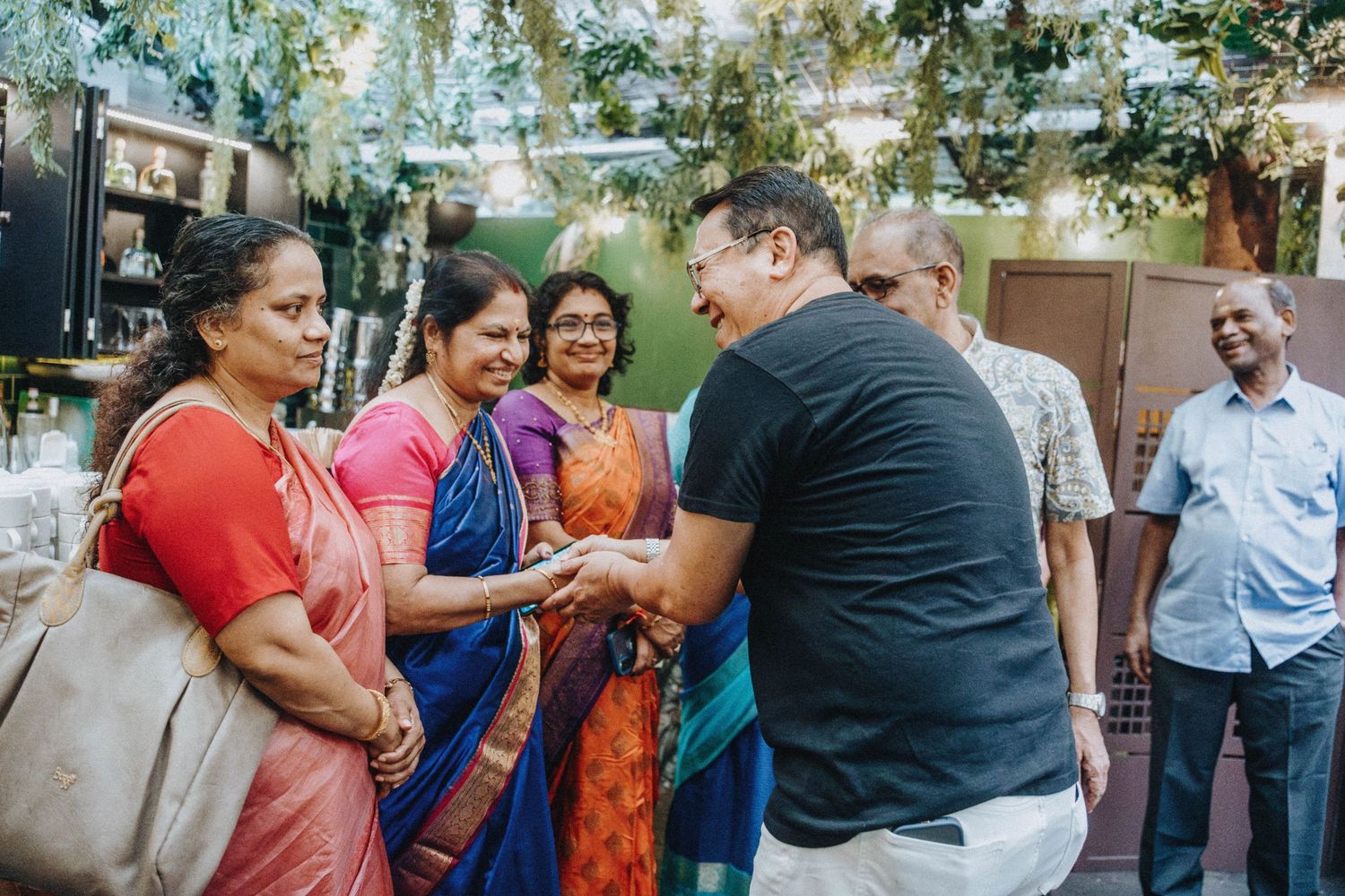 Man in black shirt warmly greets women in colorful sarees at a festive outdoor gathering.