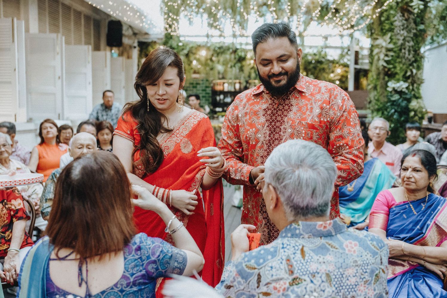 Couple in vibrant Indian wedding attire greeting elderly guests during a colorful outdoor cultural ceremony.