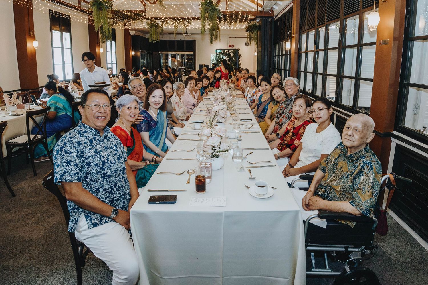 Large group of smiling wedding guests seated at a long banquet table during indoor reception dinner.