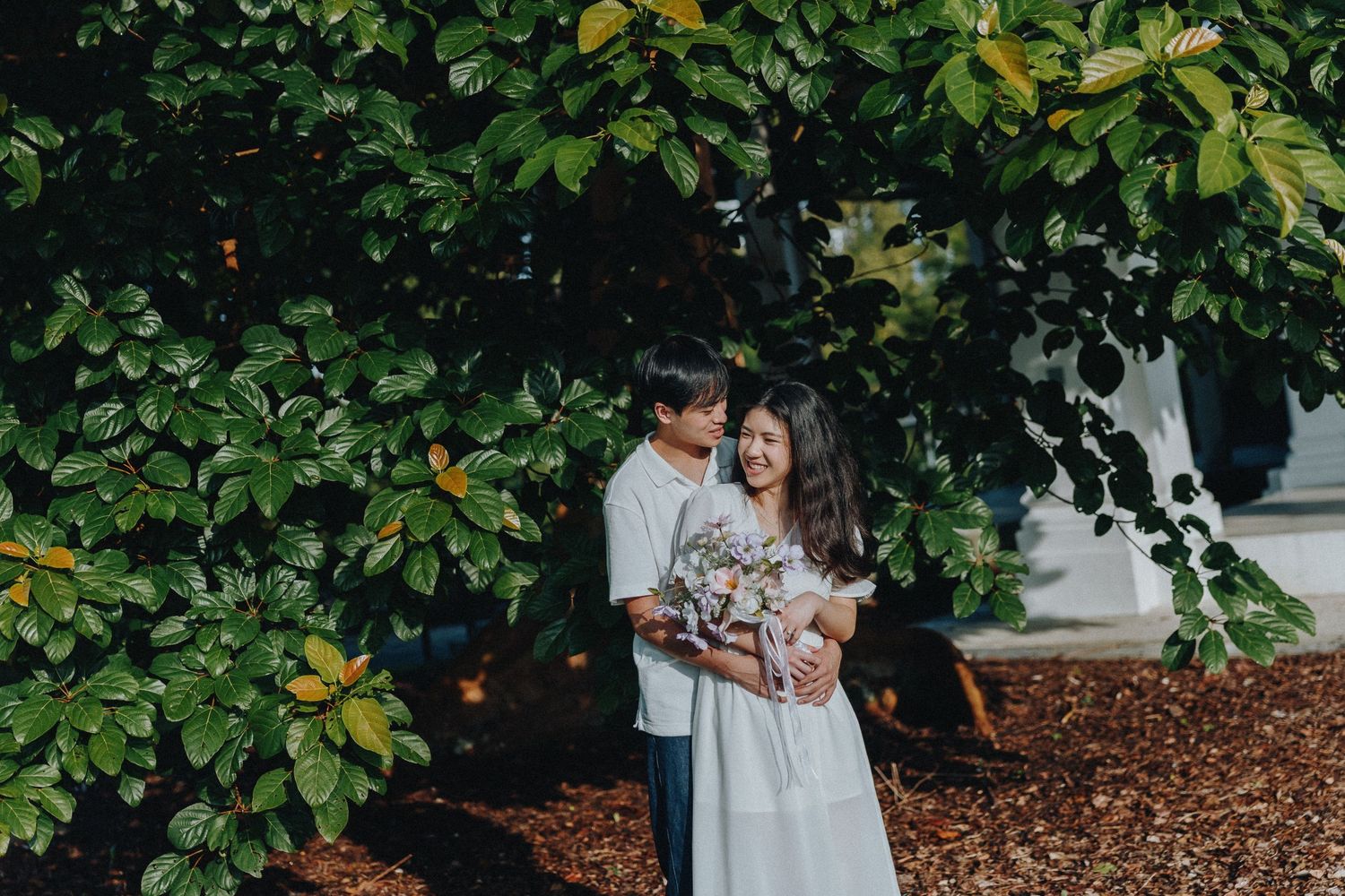 A couple stands among lush green foliage in a romantic outdoor setting.