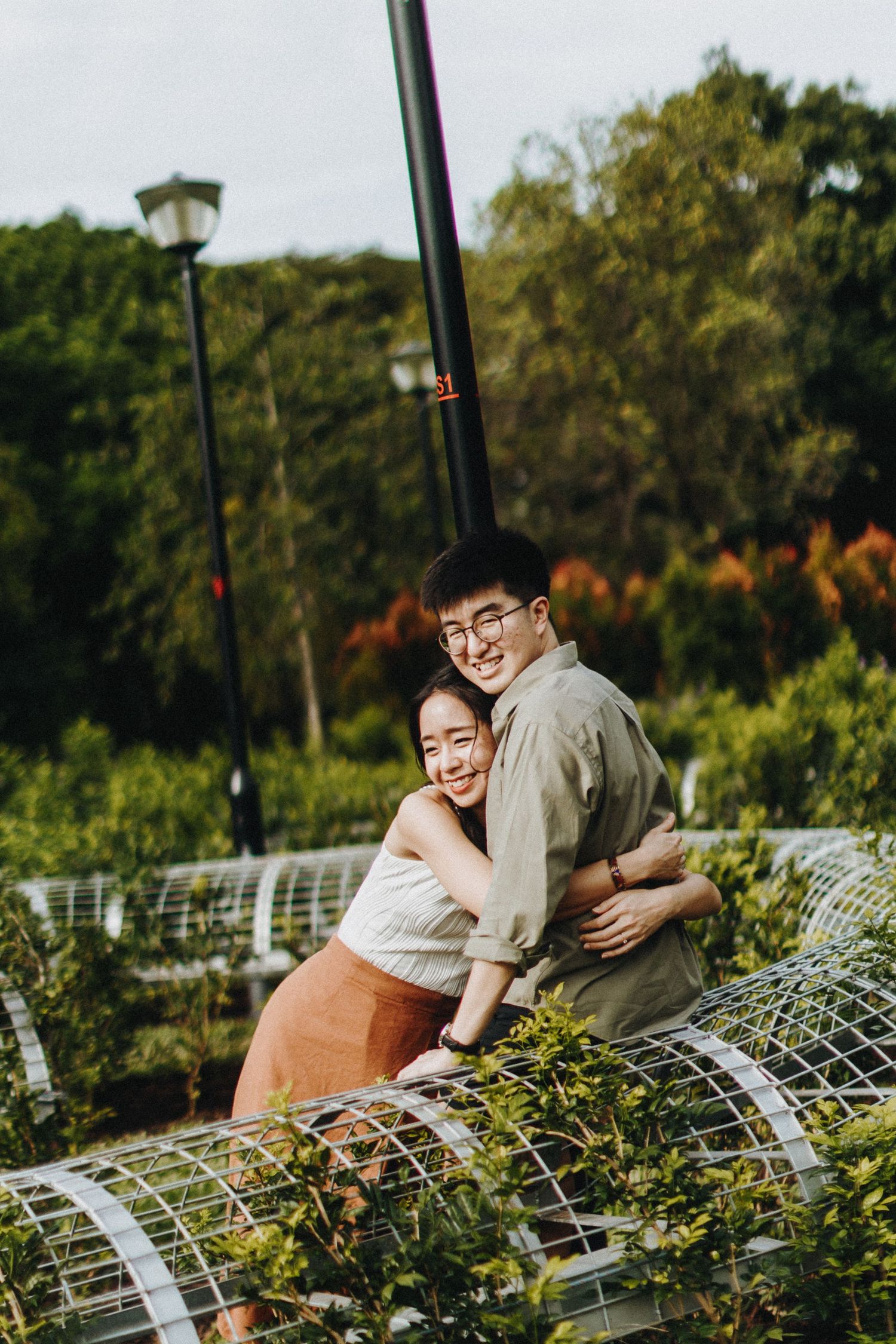 A couple embraces while leaning on garden trellises surrounded by green foliage and lamp posts.