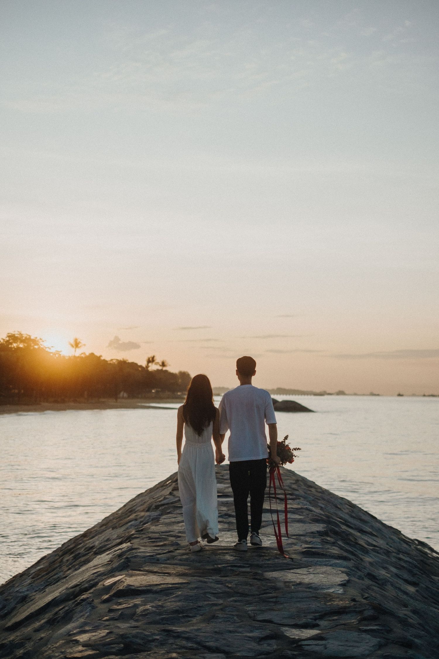 Couple walking hand in hand on stone pier during golden hour sunset.