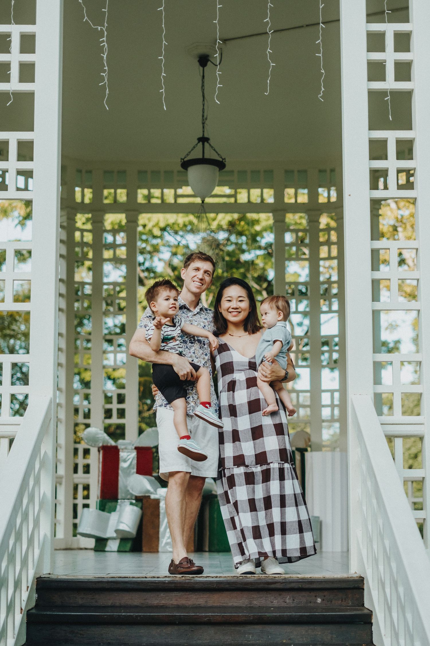 A family poses together on a white porch with lattice work and hanging lights overhead.
