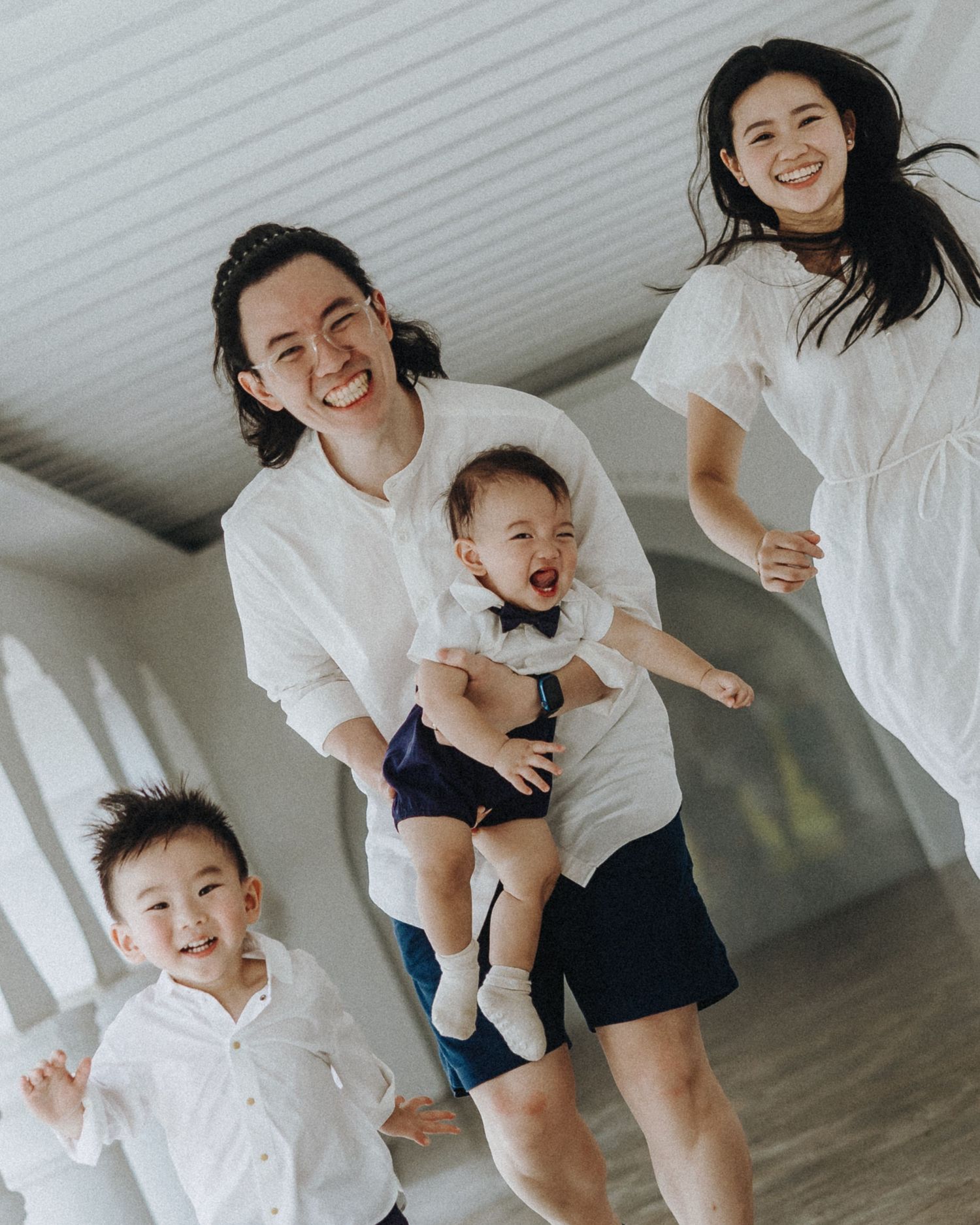 Family wearing coordinated white outfits laughing and jumping together in a bright indoor space.