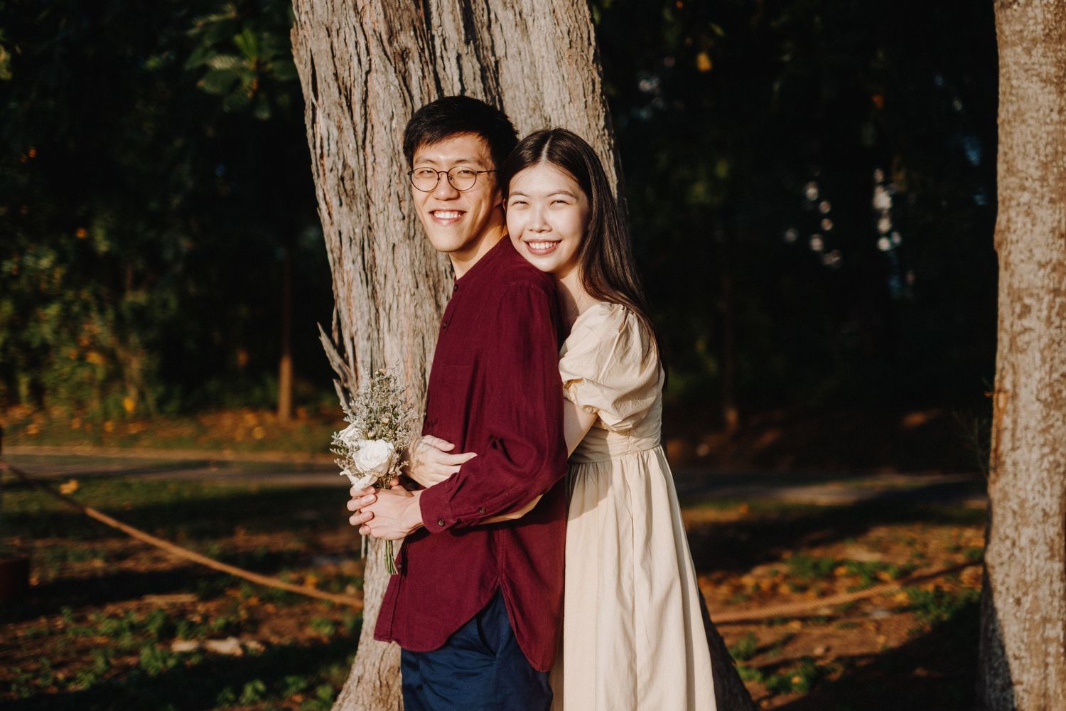 A couple embraces while standing against a tree trunk in a forest setting with autumn leaves on the ground.