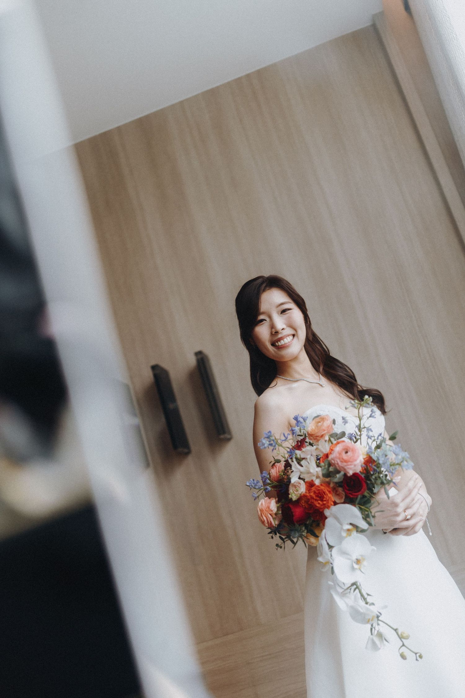 A bride in an off-shoulder wedding gown holding a cascading bouquet of pink and coral flowers against a wooden wall.