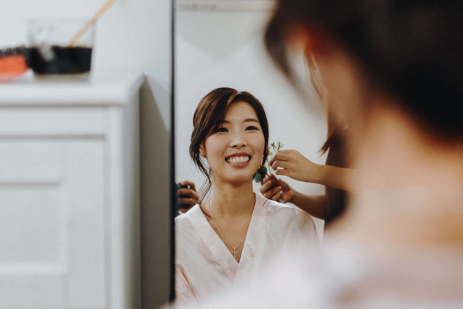 Stylist adjusts earrings on client during wedding preparation in bright white getting-ready room.