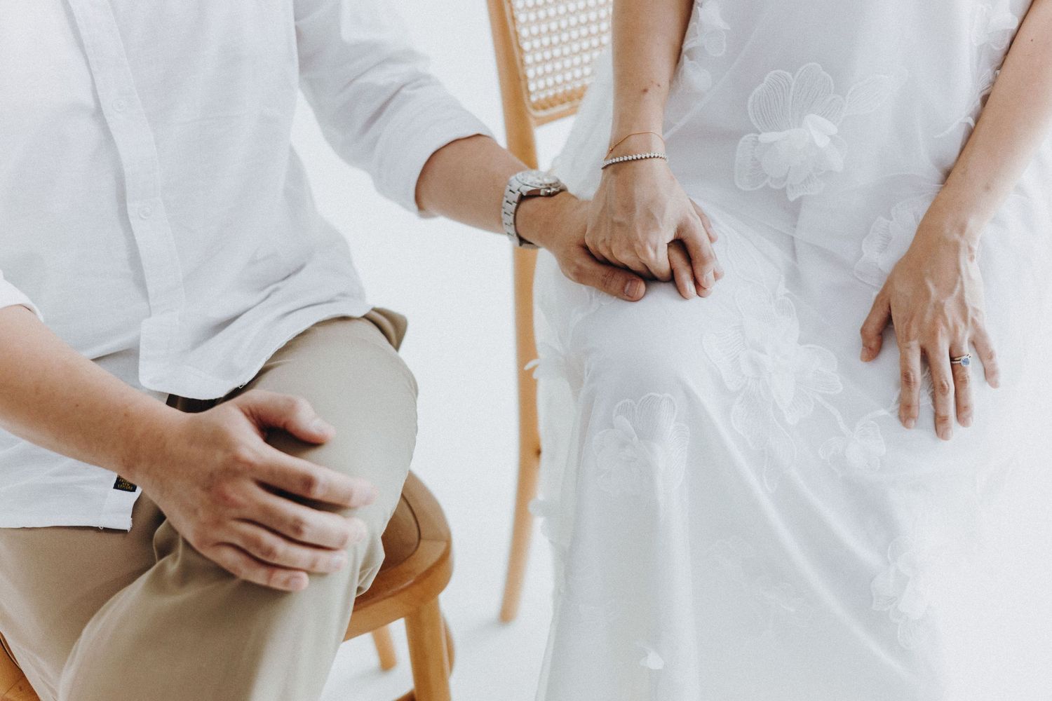 Two people in white outfits holding hands while sitting together against a light minimalist background.