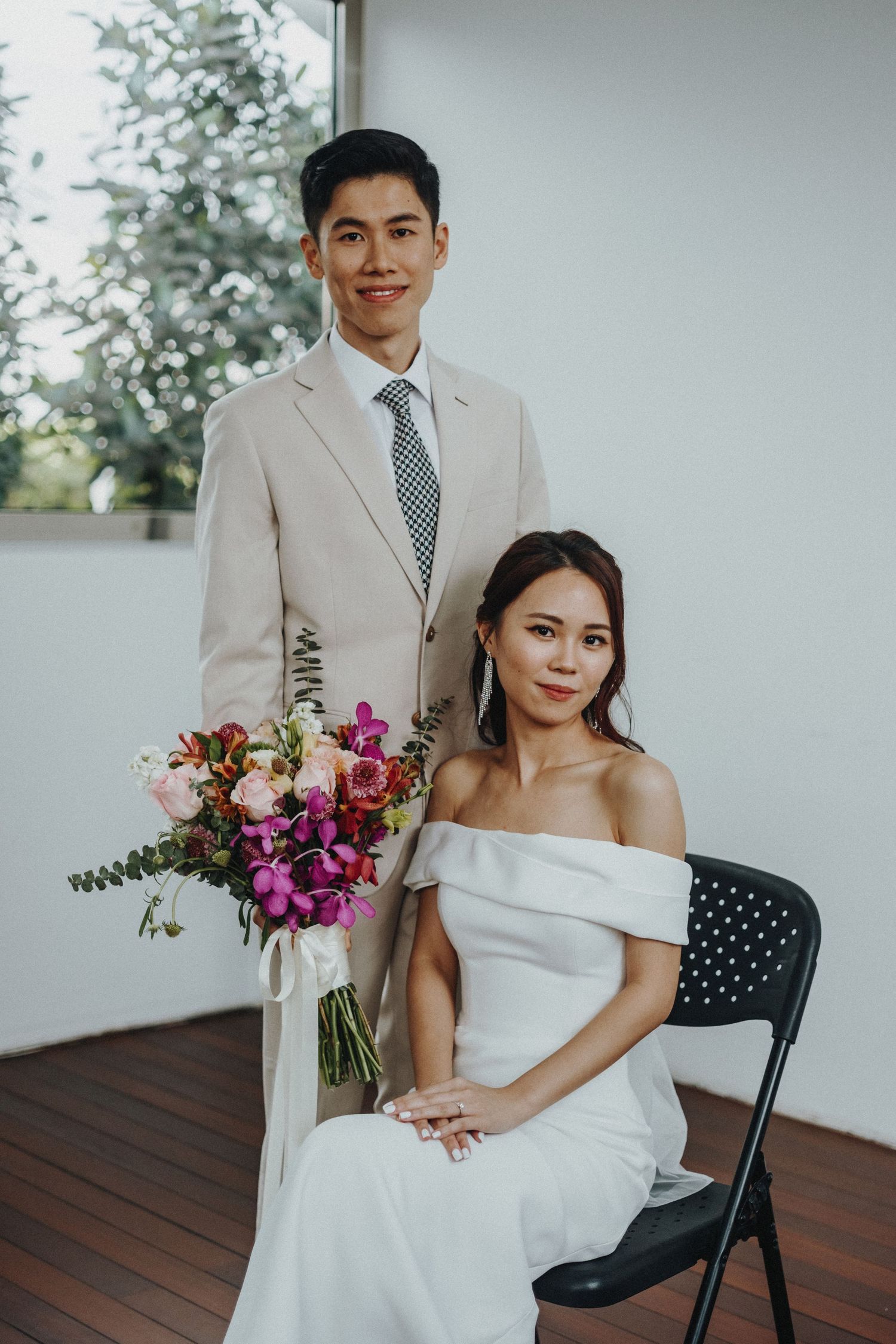 Wedding couple in formal attire pose with vibrant pink and purple floral bouquet on outdoor deck.