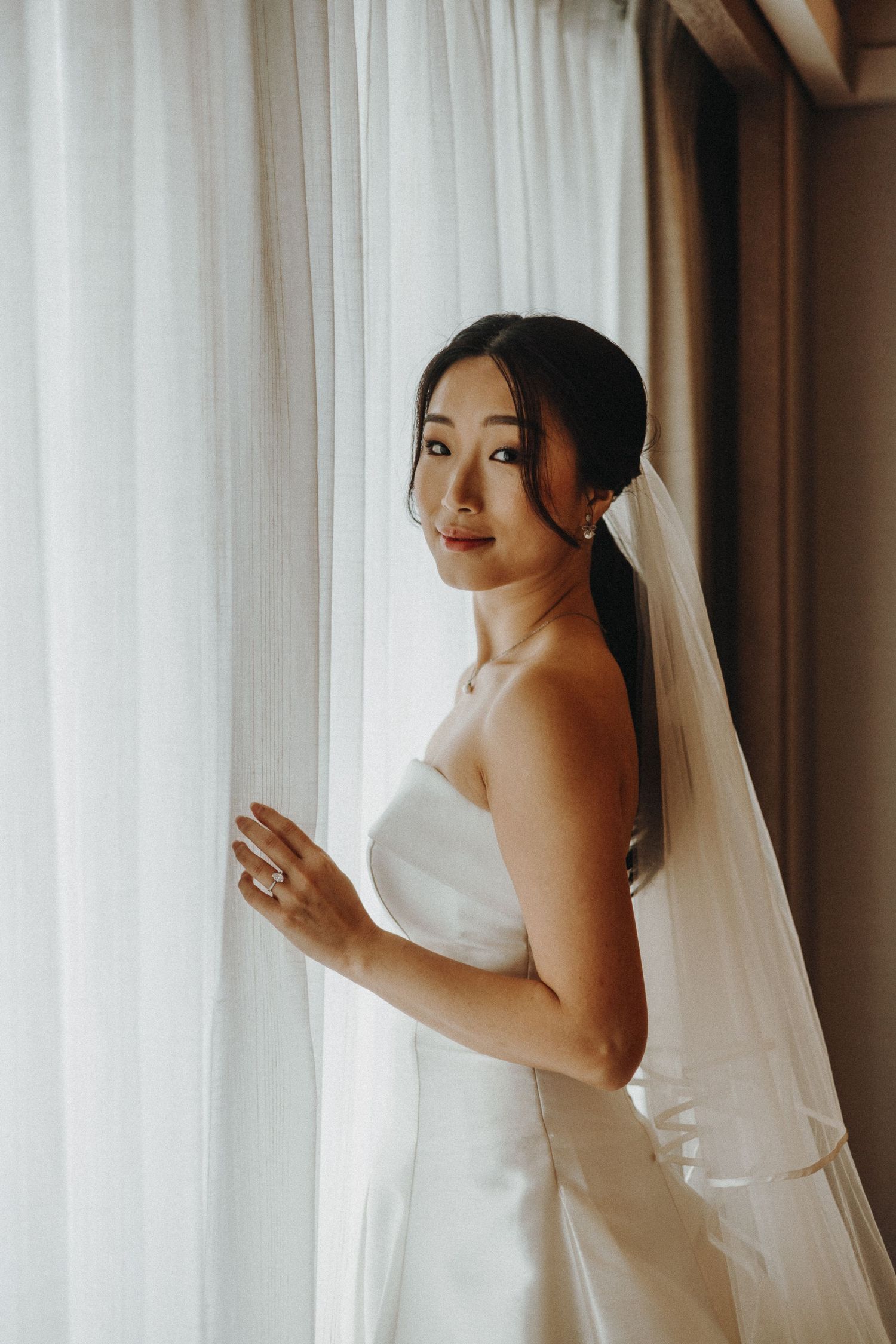 A bride in a white wedding dress and veil poses elegantly by sheer curtains in a natural light setting.