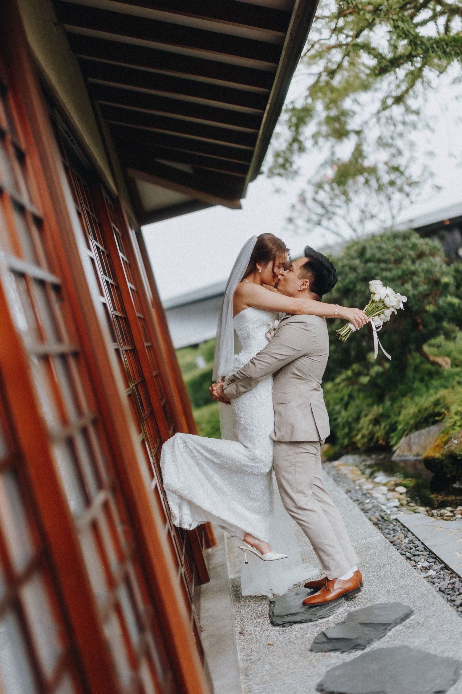 Newlyweds share romantic dance pose outside traditional Asian building.