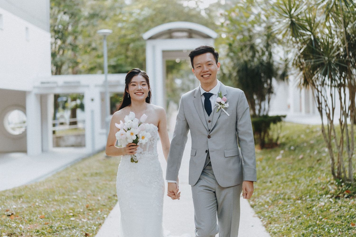 A newlywed couple walks hand in hand along a garden path surrounded by white buildings and greenery.