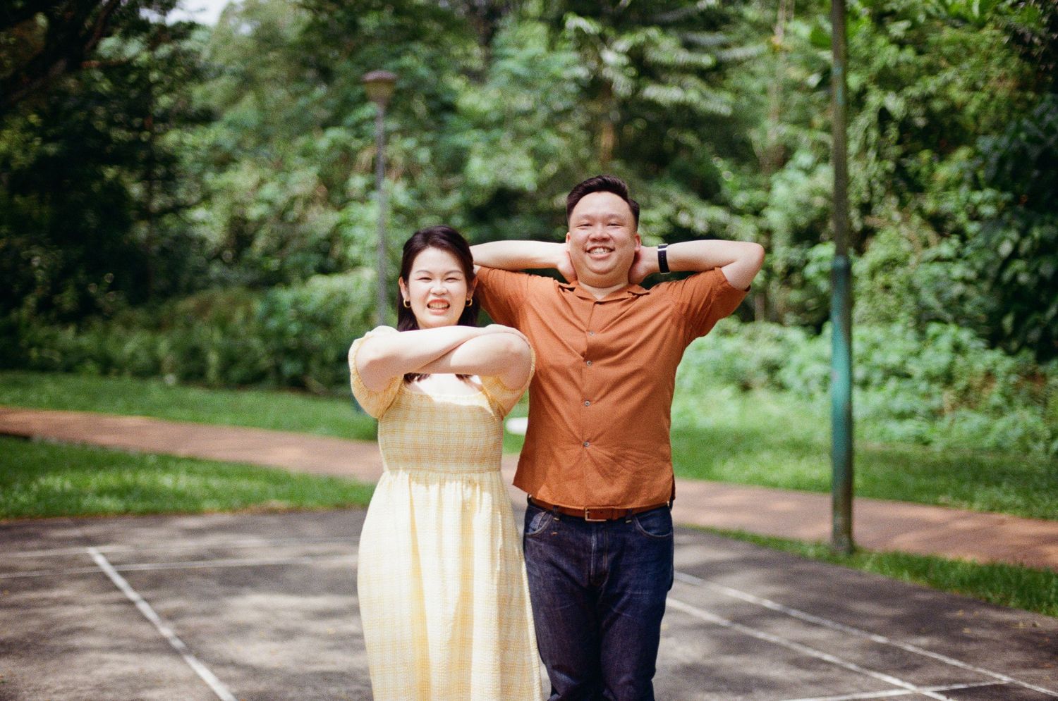 Two people in casual attire pose playfully with arms up on a walking path surrounded by greenery.