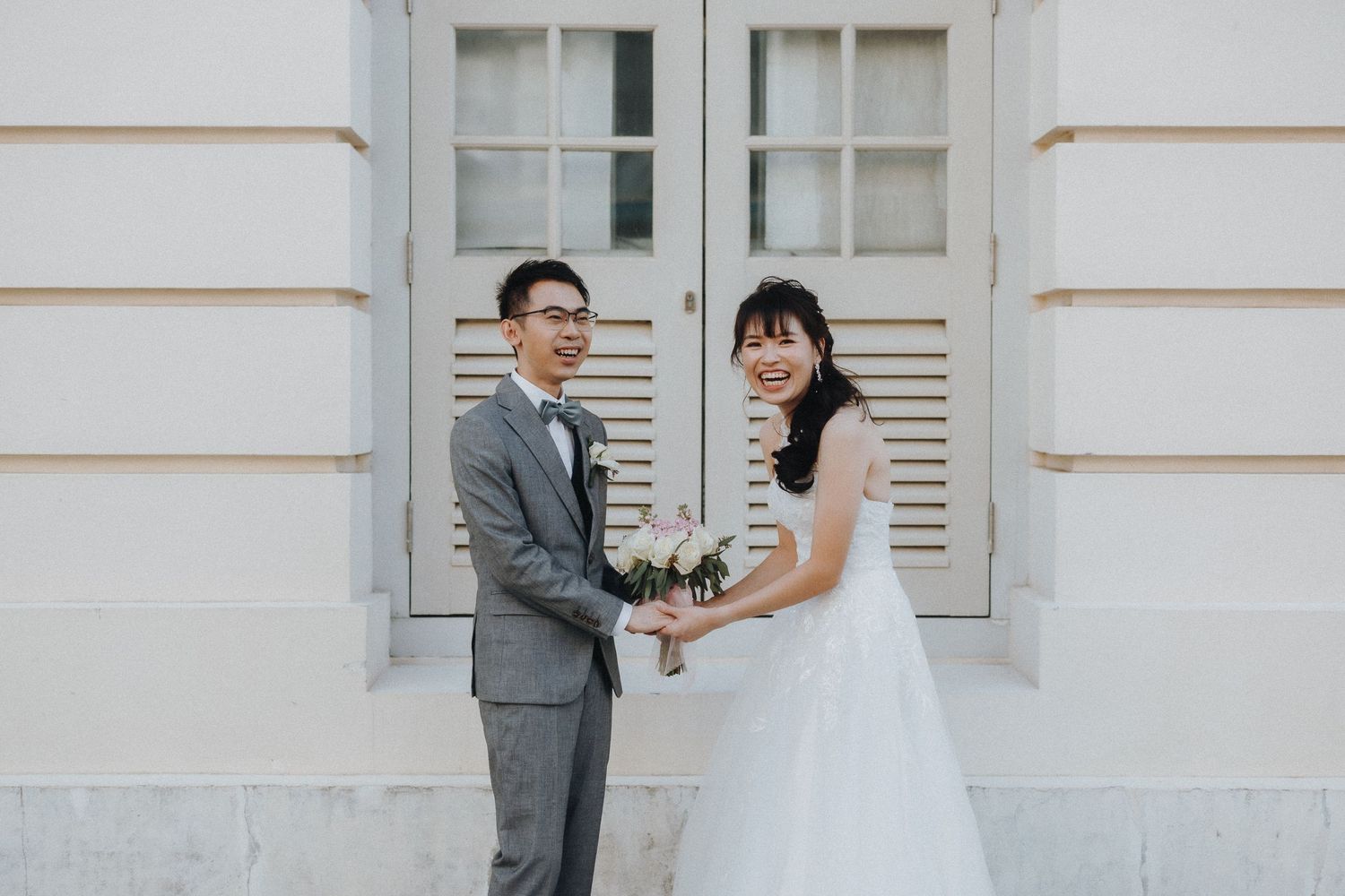 A wedding couple poses in front of white shuttered doors wearing traditional formal attire.