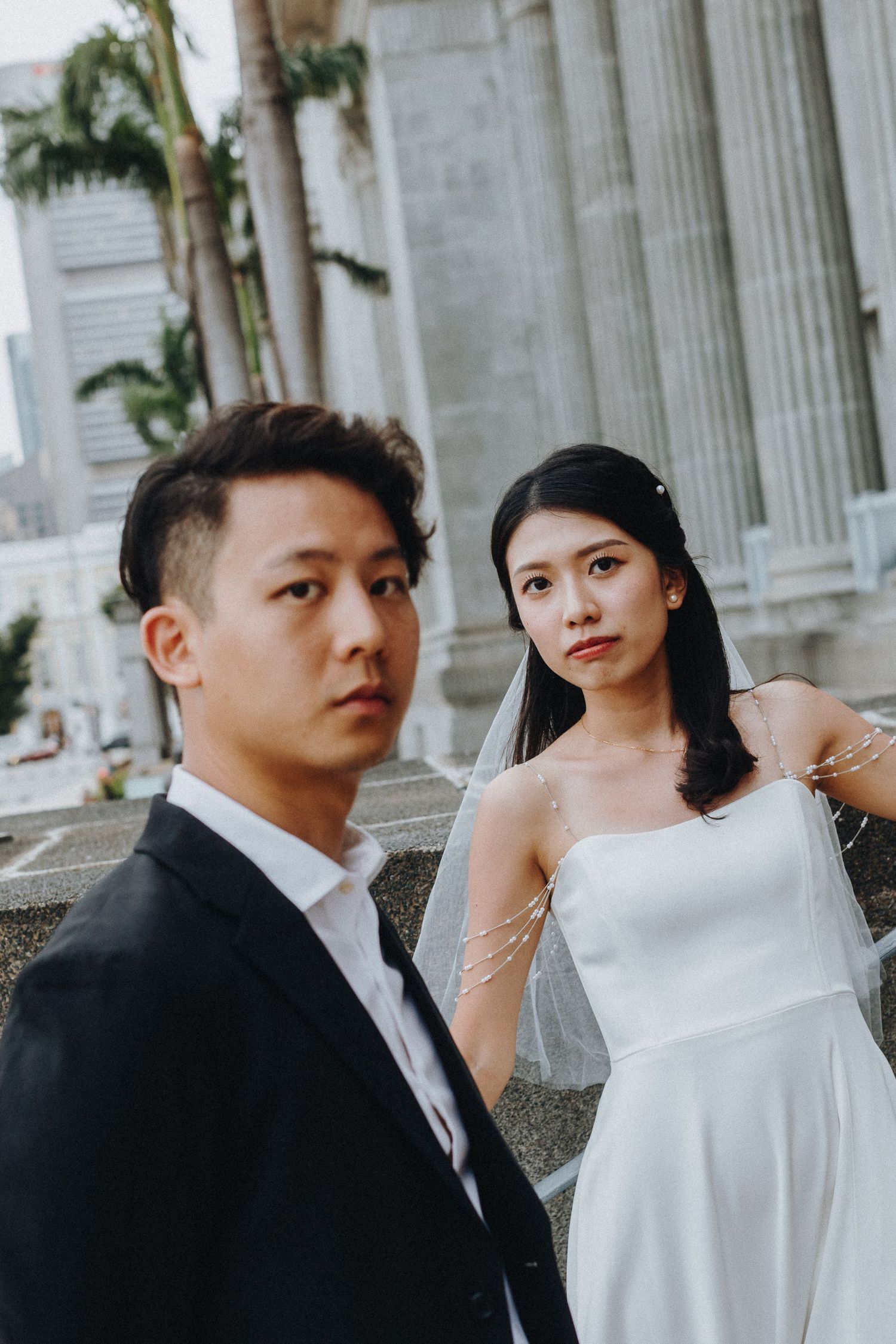A couple in formal wedding attire poses in front of classical stone columns and architecture.