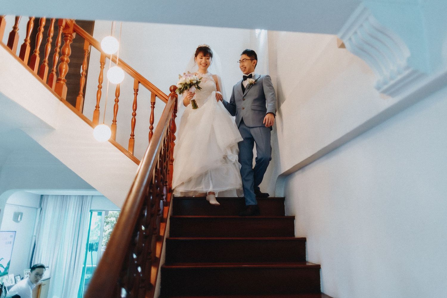 A couple descending wooden stairs with white walls, bride holding a bouquet of flowers.