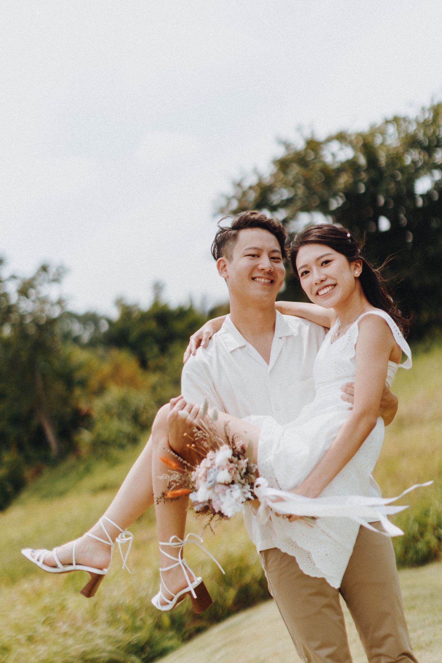 A playful wedding photoshoot in a sunny field with the couple in white and khaki attire sharing joyful moments.