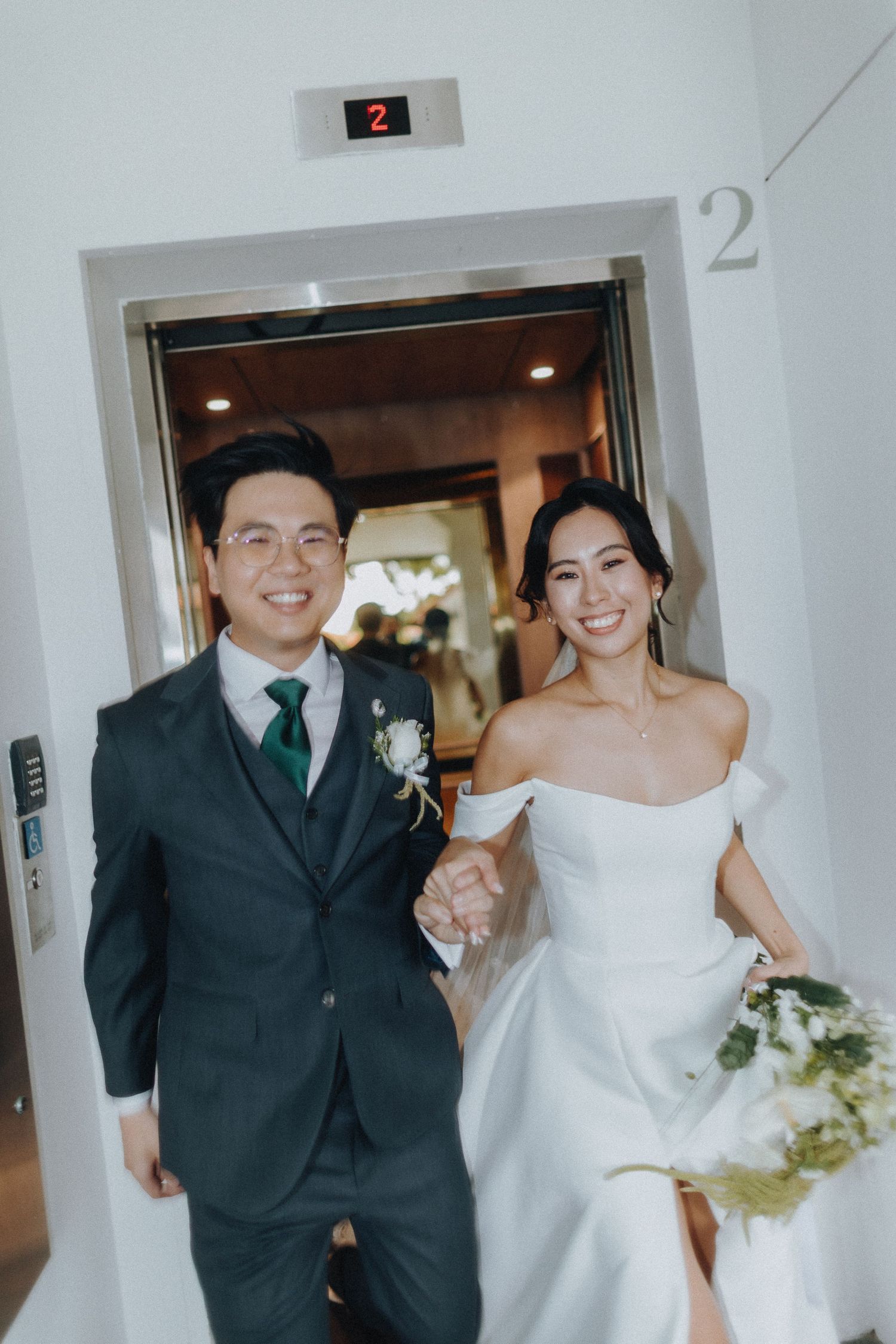 A wedding couple steps out of an elevator into a hallway, the bride in white and groom in dark suit with green tie.