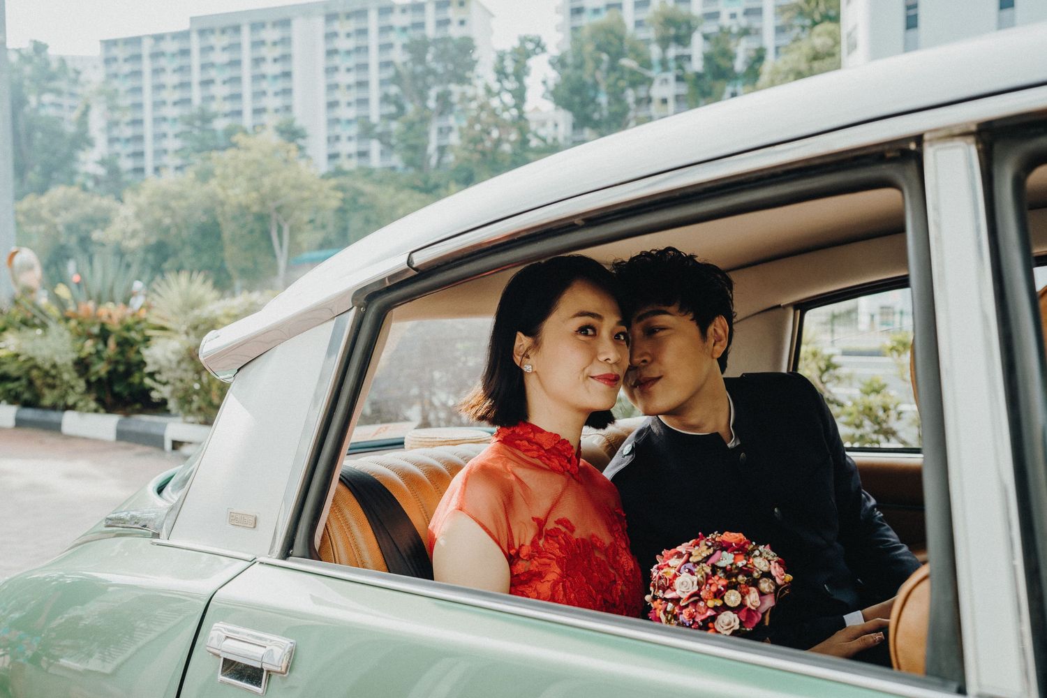 Couple sharing intimate moment in vintage car with red dress and bouquet.