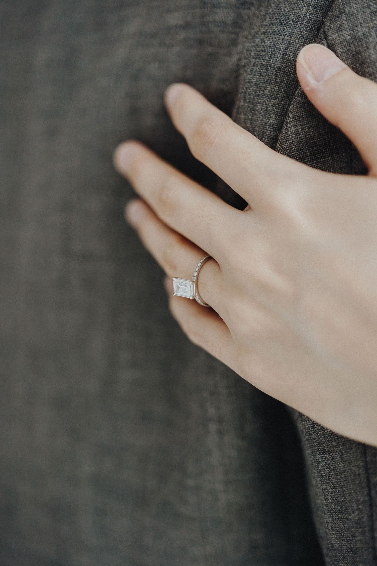 Close-up view of an elegant diamond engagement ring on a hand against dark gray fabric.