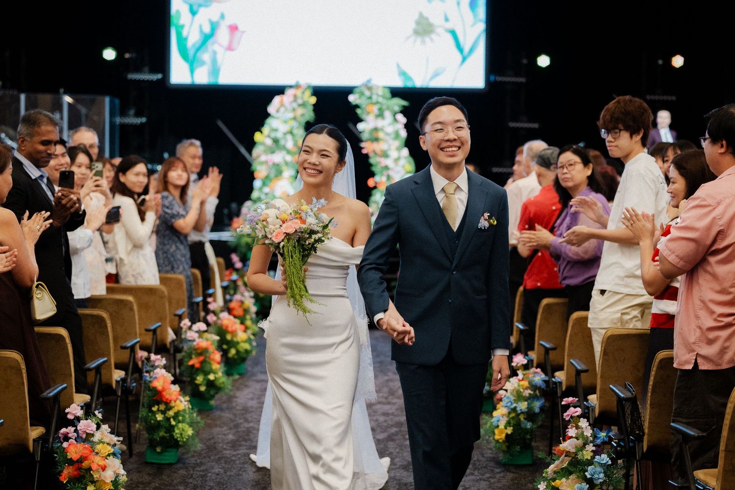Newlywed Asian couple walks joyfully down the aisle hand in hand after wedding ceremony with guests applauding.