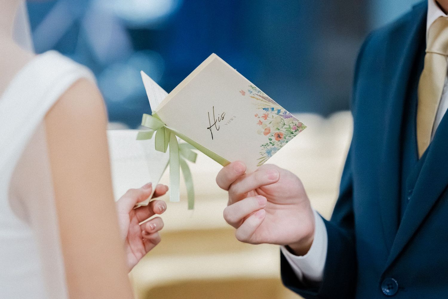 Close-up of bride and groom holding a floral-decorated vow booklet tied with a green ribbon during ceremony.