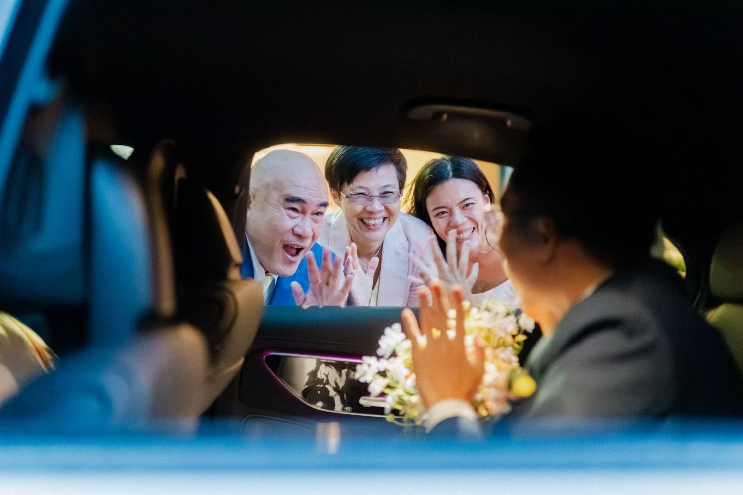 Family members waving and smiling at newlyweds through a car window after wedding.