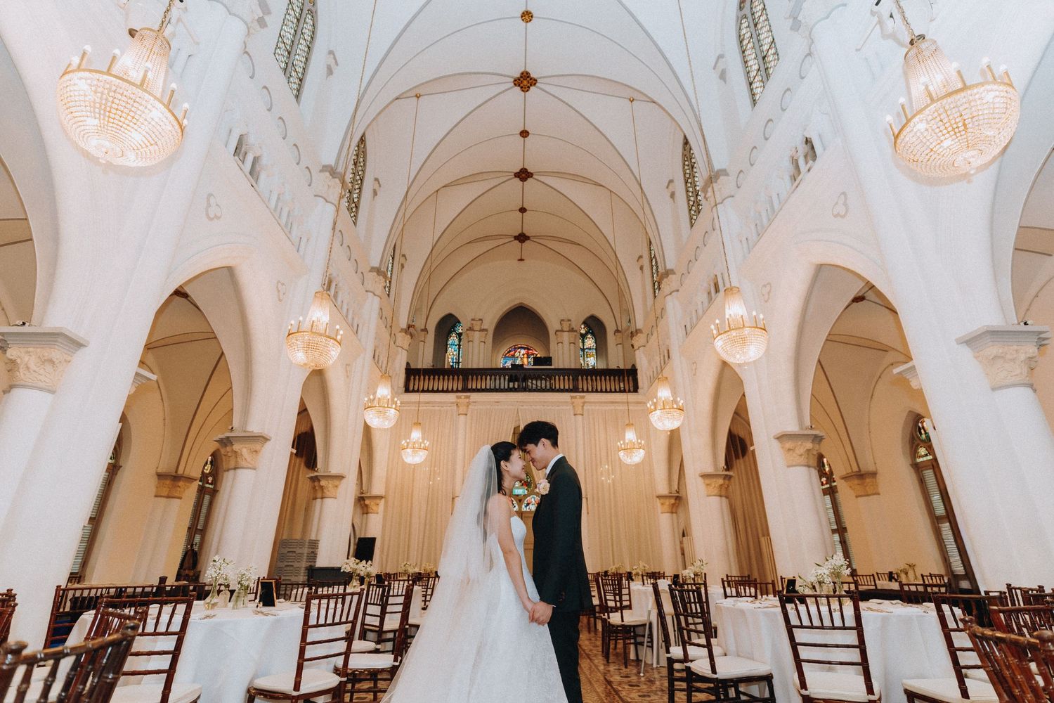 Bride and groom share a romantic kiss inside an elegant white church with arched ceilings and chandelier lighting.