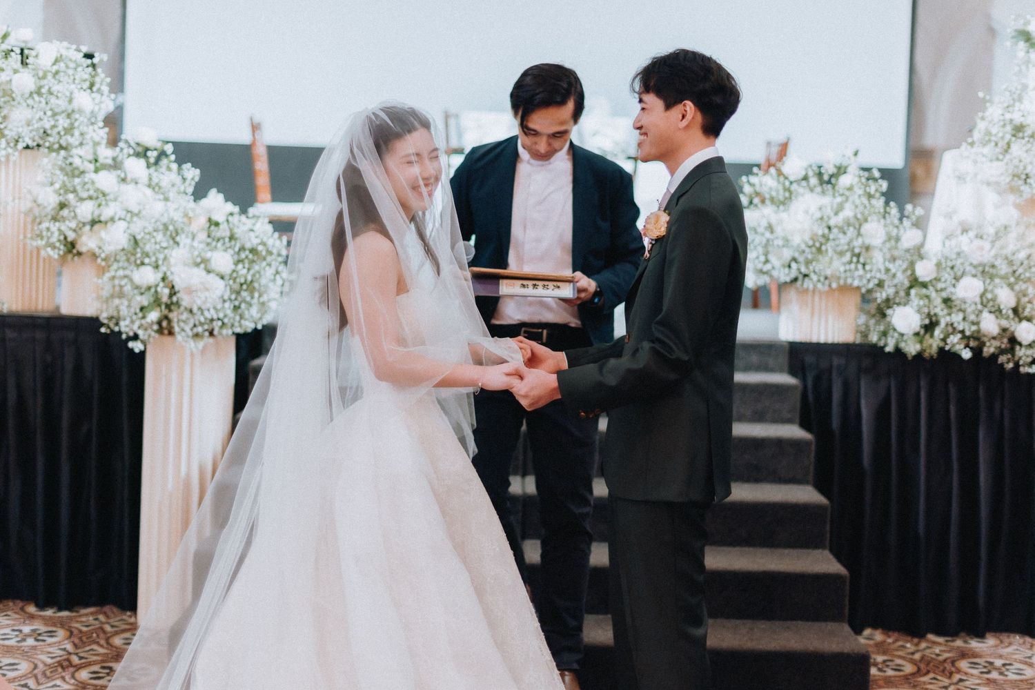 Bride and groom exchanging rings at elegant wedding ceremony with white floral arrangements and officiant.