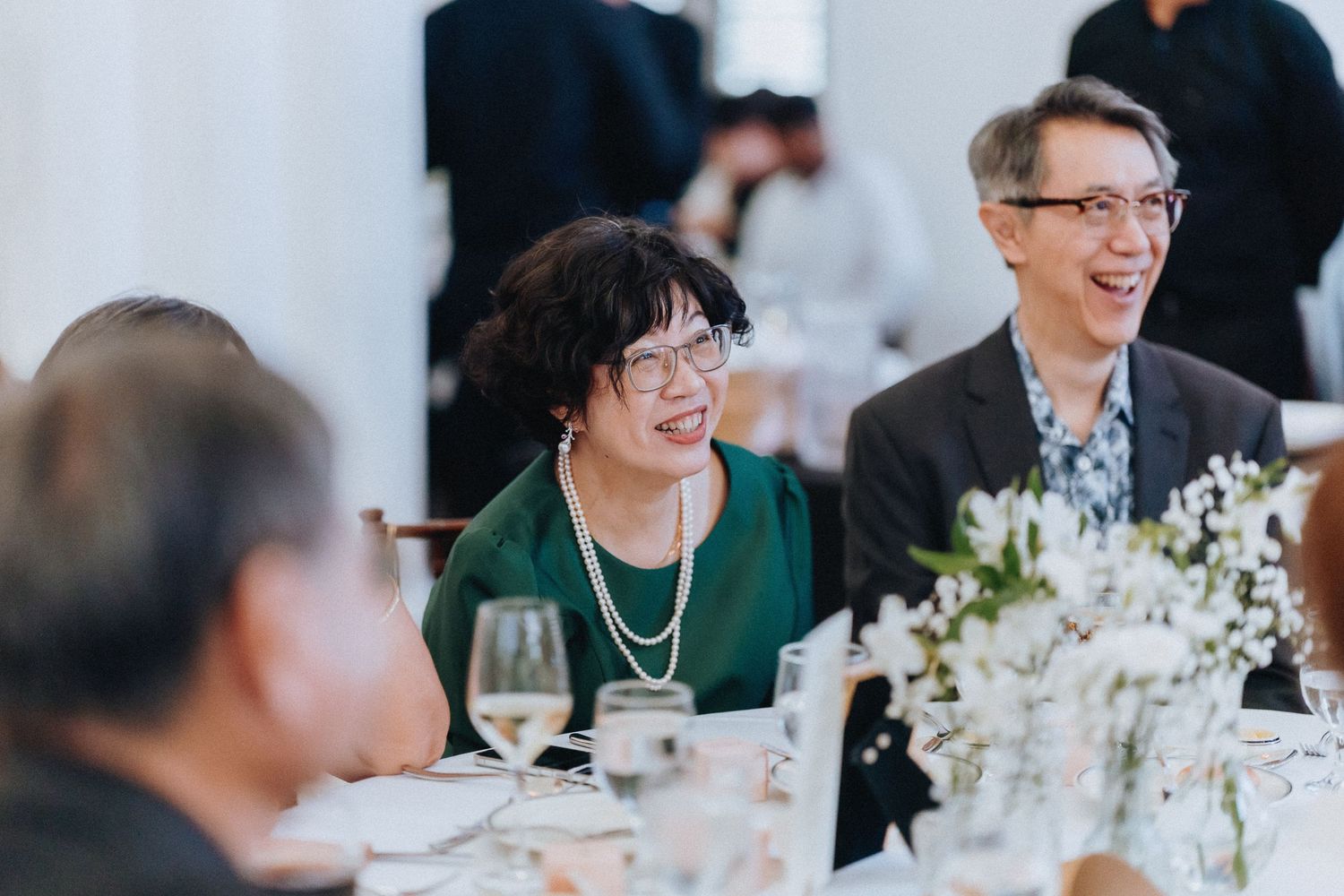 Two smiling wedding guests, a woman in green dress with pearls and a man in glasses, seated at a reception dinner table.