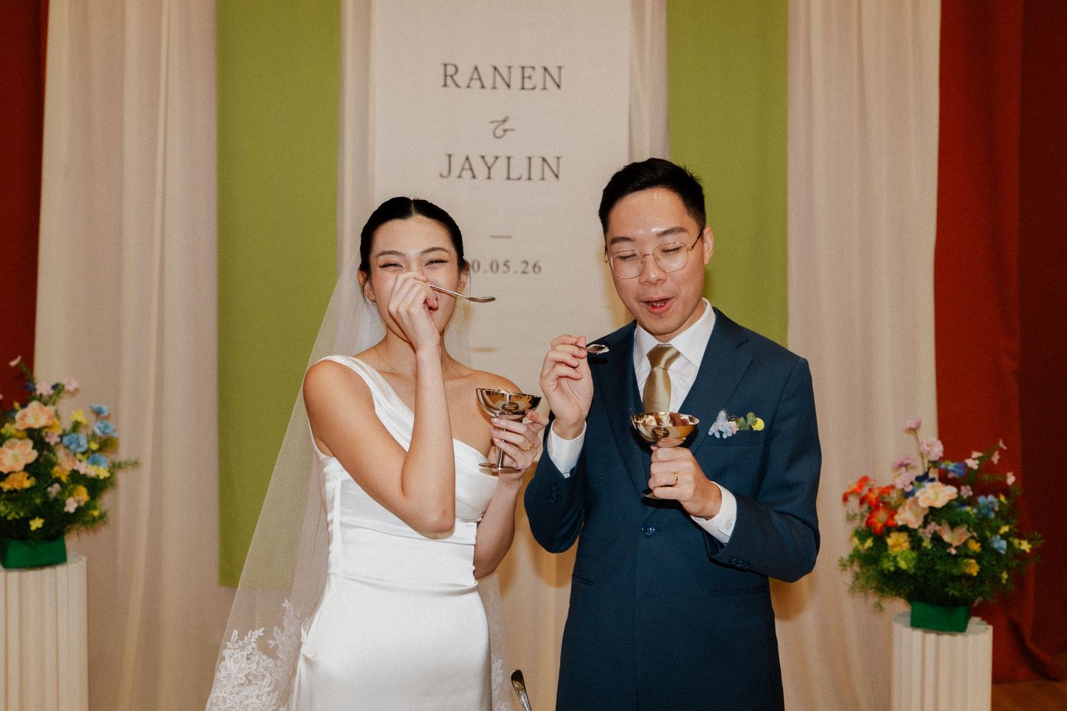 Bride and groom laughing while holding golden cups at wedding reception with floral decorations.