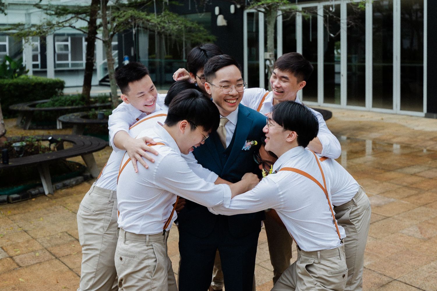 Groom in navy suit is joyfully embraced by groomsmen in white shirts during outdoor wedding celebration.