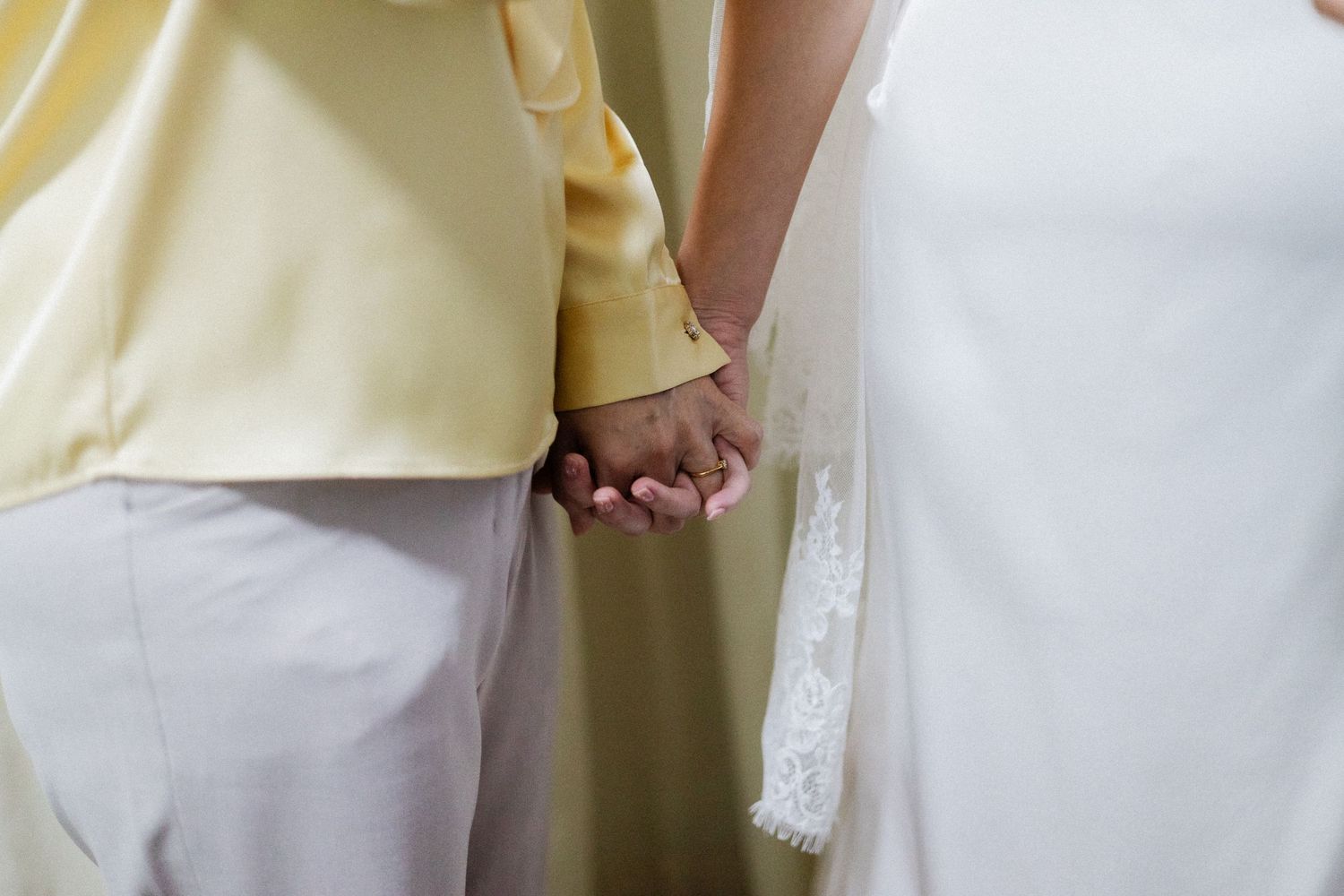 Couple holding hands in wedding attire, bride in white lace gown and groom in yellow shirt with white pants.