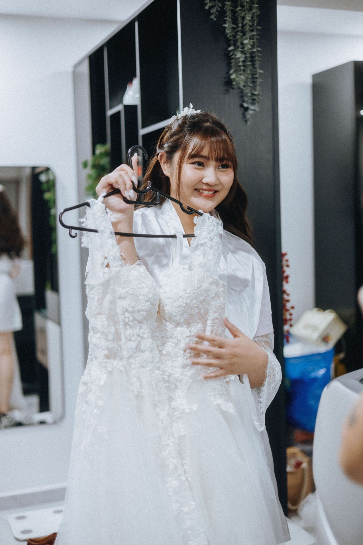 A white lace wedding dress hangs on display in a bridal boutique with hanging plants in the background.