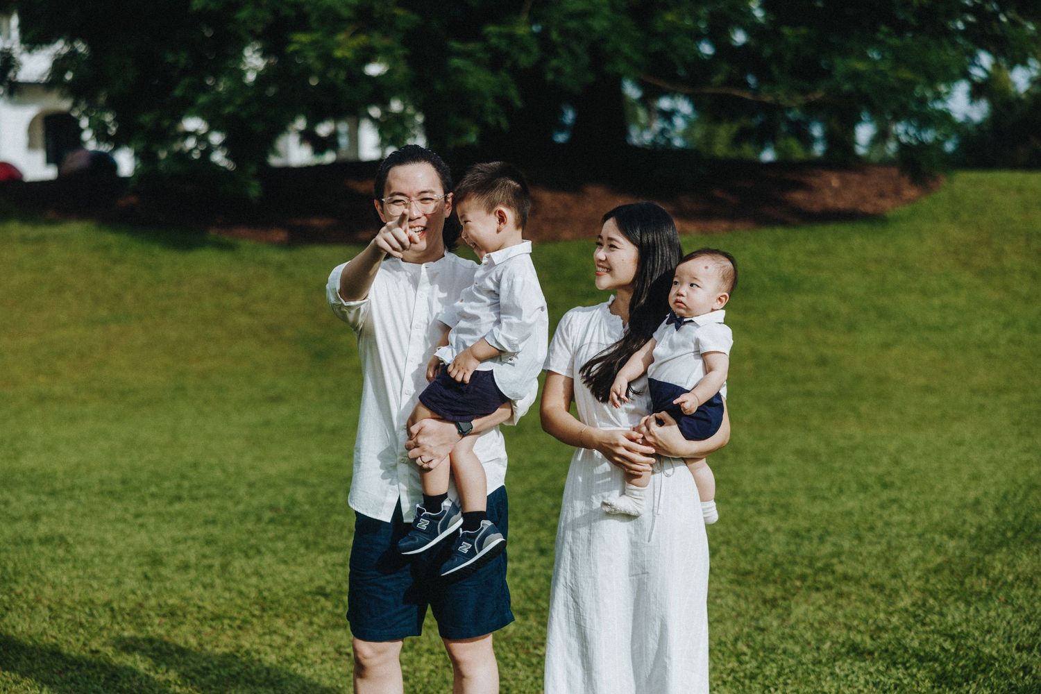 A couple stands together with their children on a grassy lawn in a park on a summer day.