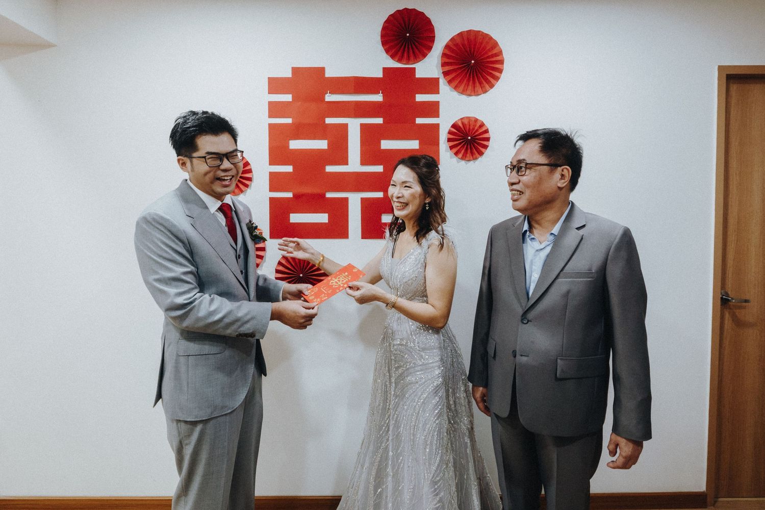 People in formal attire sharing a laugh in front of a traditional Asian wedding backdrop with red lettering.