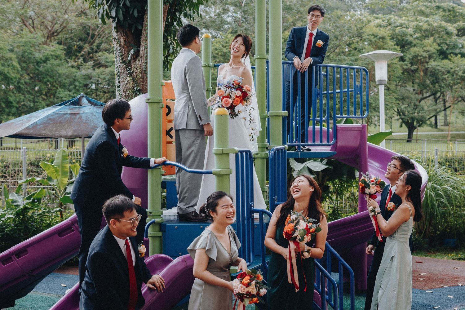 Bridesmaids and groomsmen gather on a playful purple and green playground structure for wedding photos.