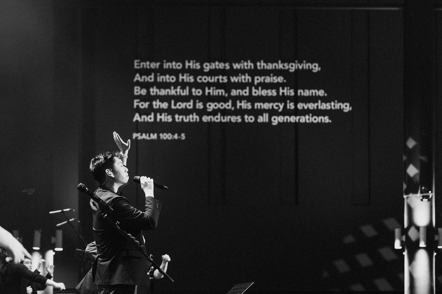 Black and white concert photo with Bible verse from Psalm 100:4 displayed over a silhouetted performer on stage.