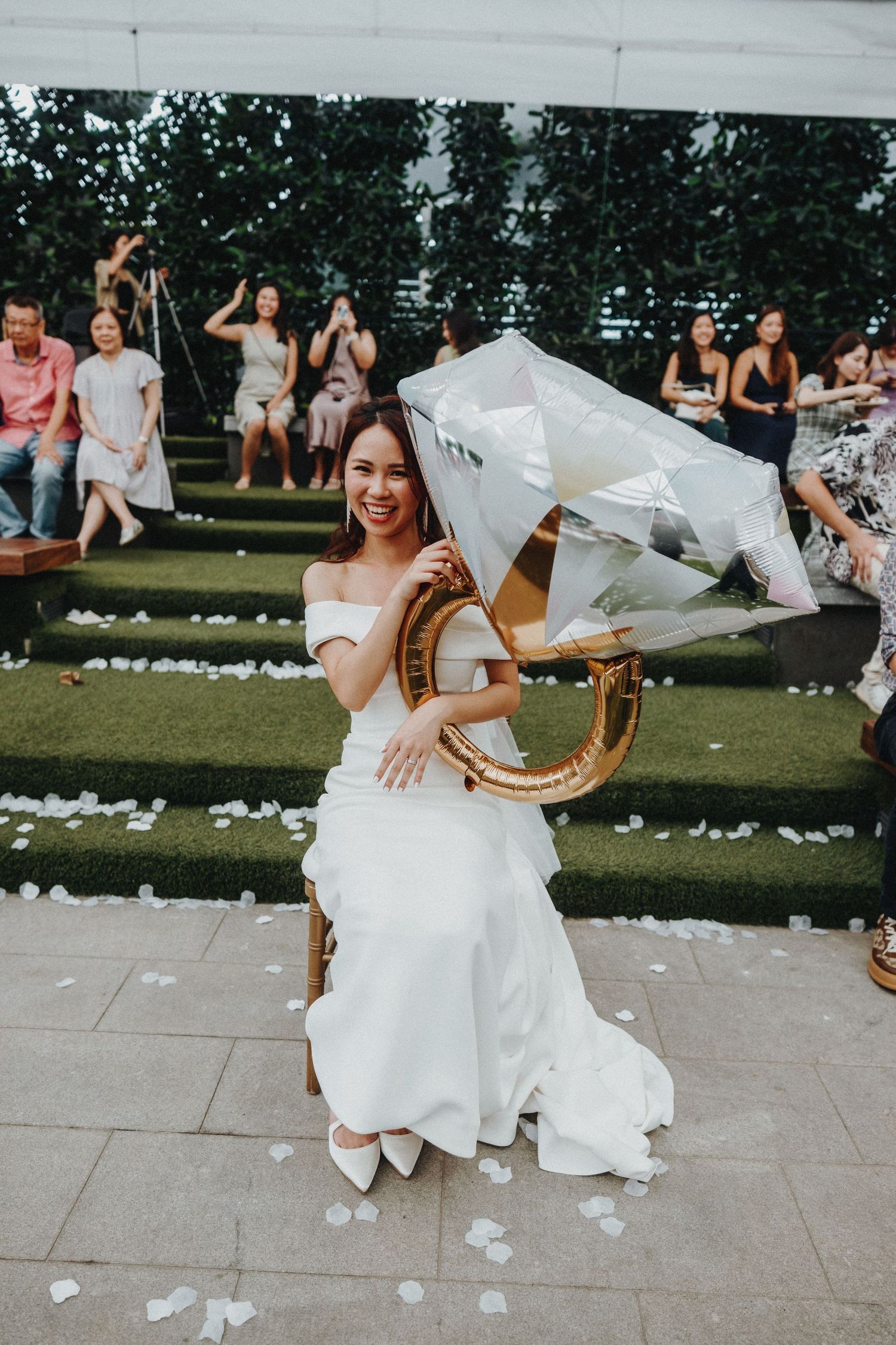 A bride in white holds a gold decorative prop during an outdoor wedding celebration.
