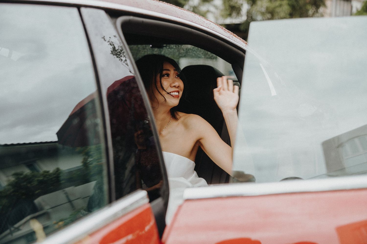 Happy bride in white dress sitting in red vintage car smiles through window.