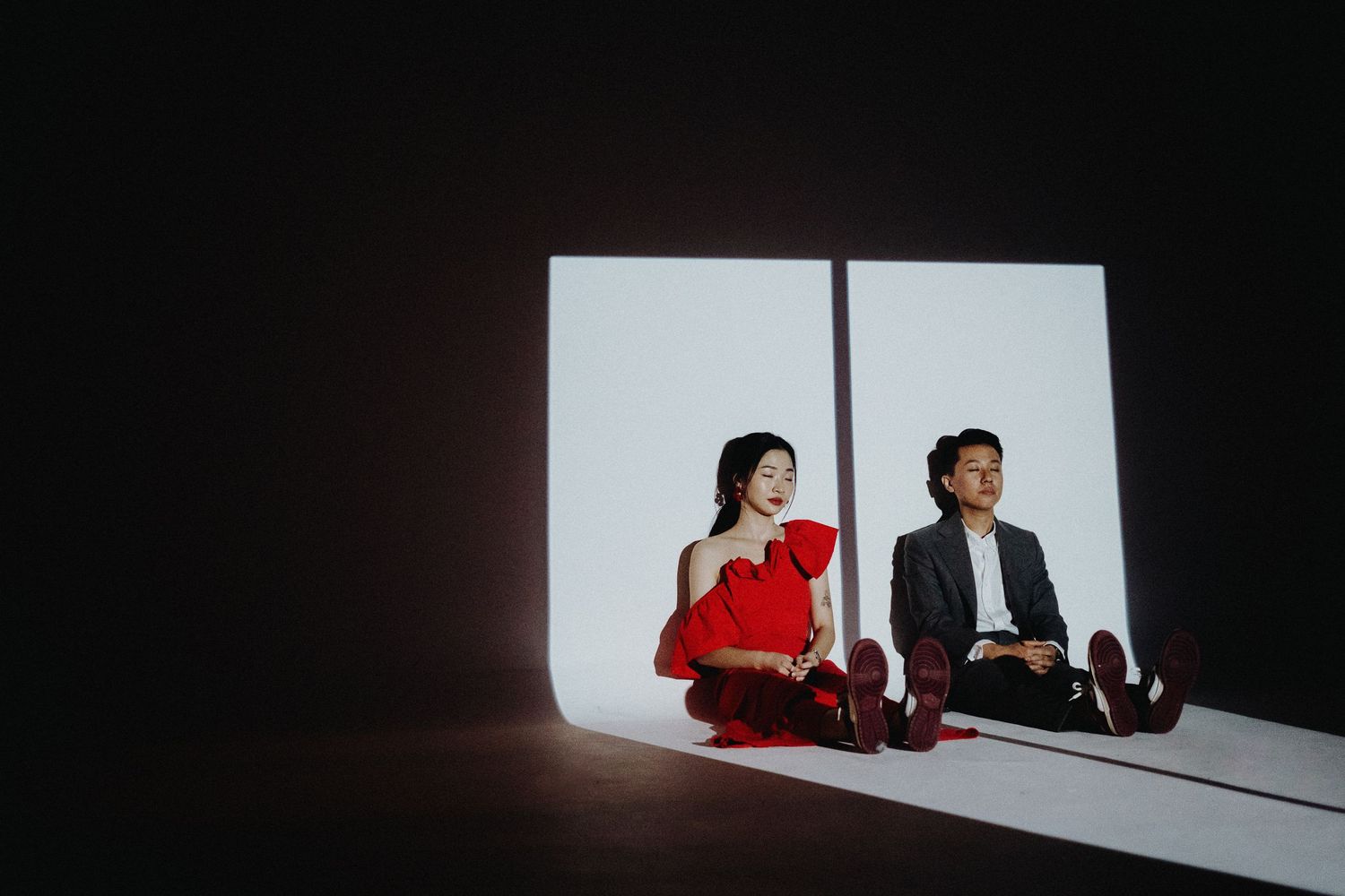 Two people in black and red sit against a white wall casting dramatic shadows in a minimalist setting.