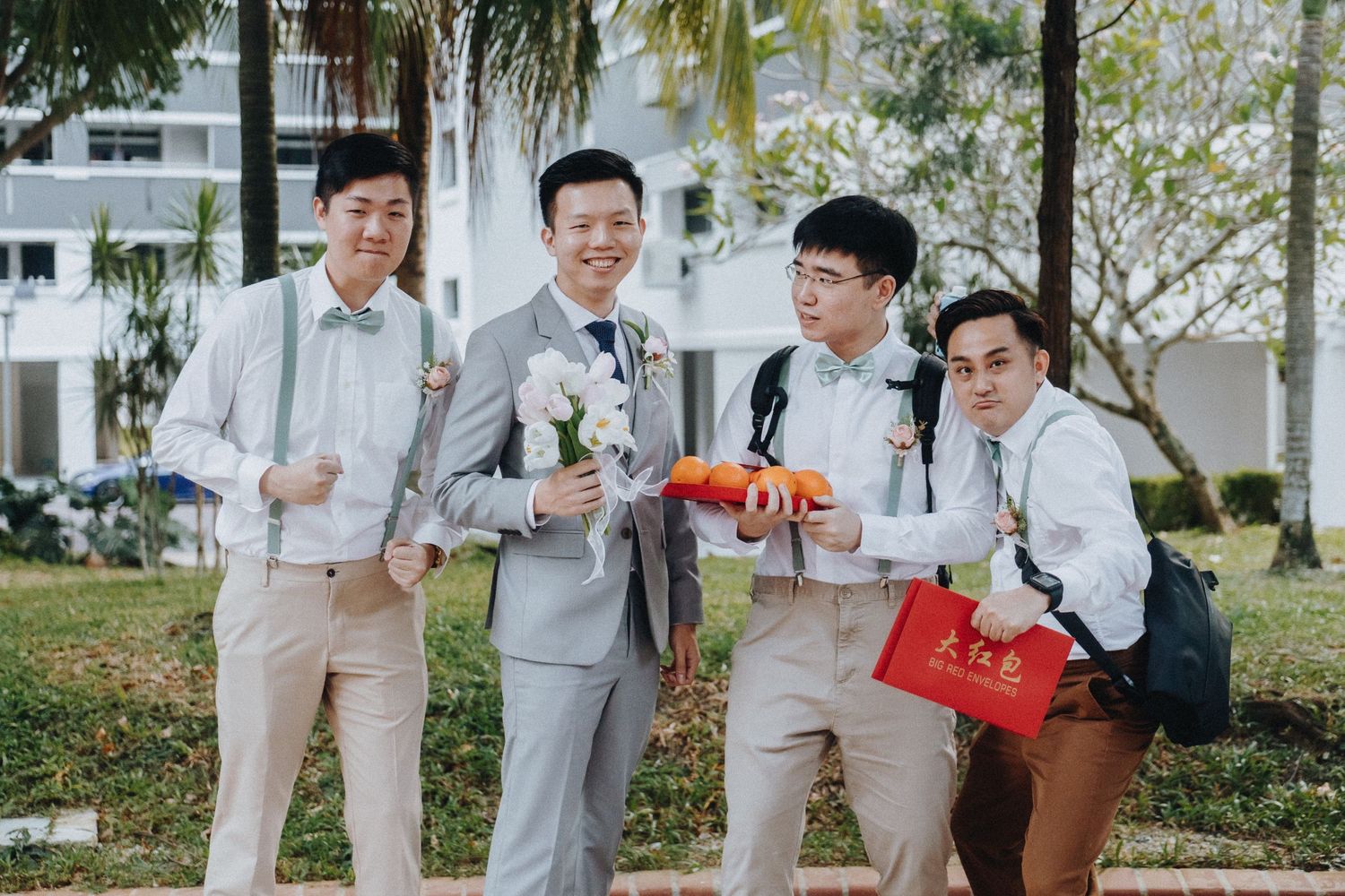 A group of groomsmen in light colored suits pose playfully outdoors with red wedding gifts.