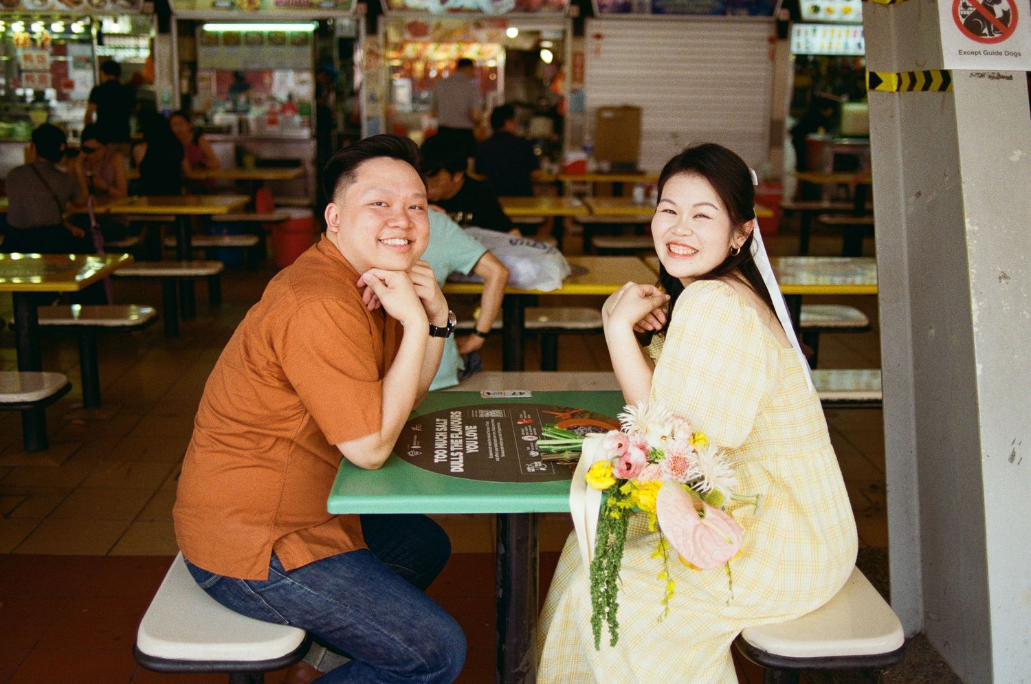A couple sits at a food court table while holding flowers in a casual dining setting.