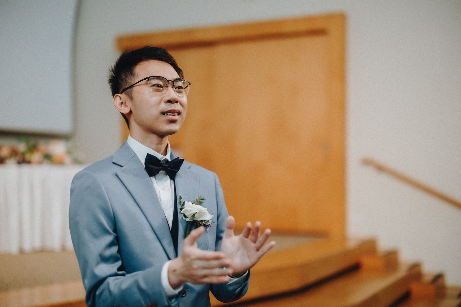 A person in a light grey suit and bowtie speaks while making gestures with their hands.