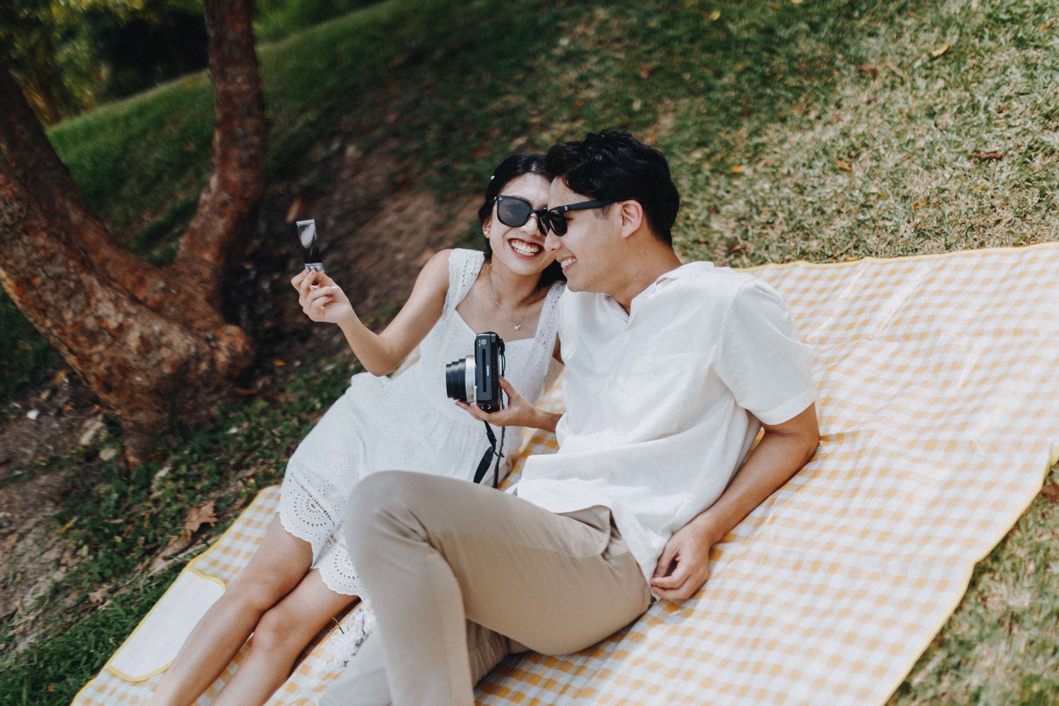 Couple relaxing on picnic blanket with drinks under tree in outdoor setting.