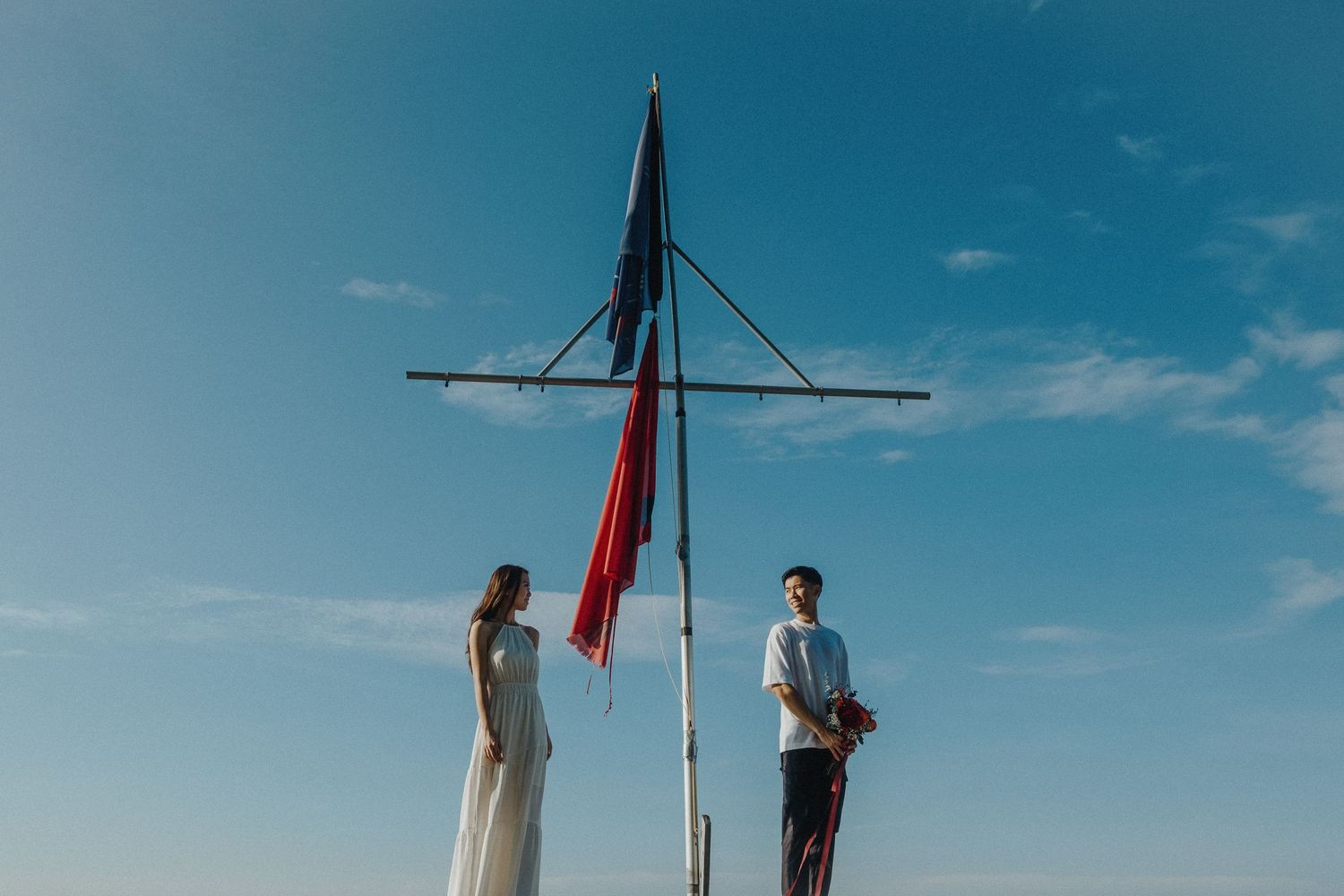 Two people stand beneath a tall flagpole against a bright blue sky.