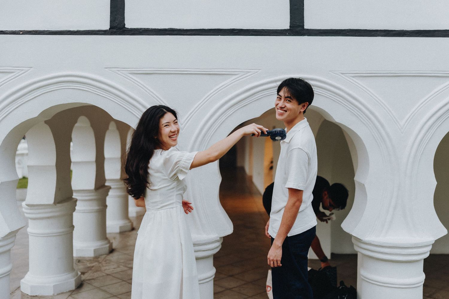 A couple in elegant wedding attire shares romantic moments under white arched columns and shadows.