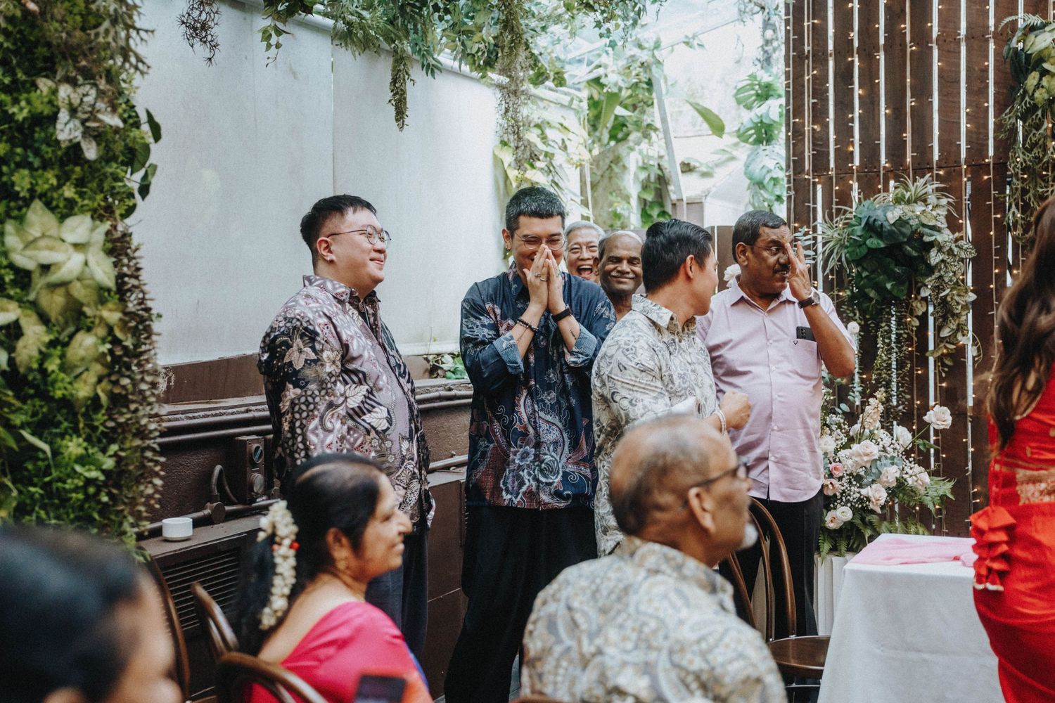 Men in batik shirts stand with hands clasped in respect at a multicultural wedding ceremony.