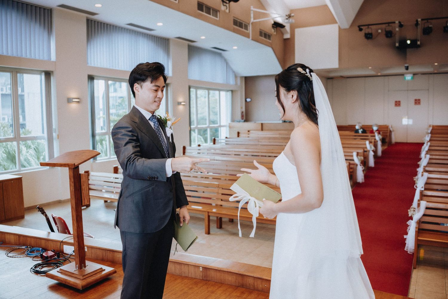 A bride and groom exchange rings during an intimate wedding ceremony inside a elegant church with red-carpeted aisles.