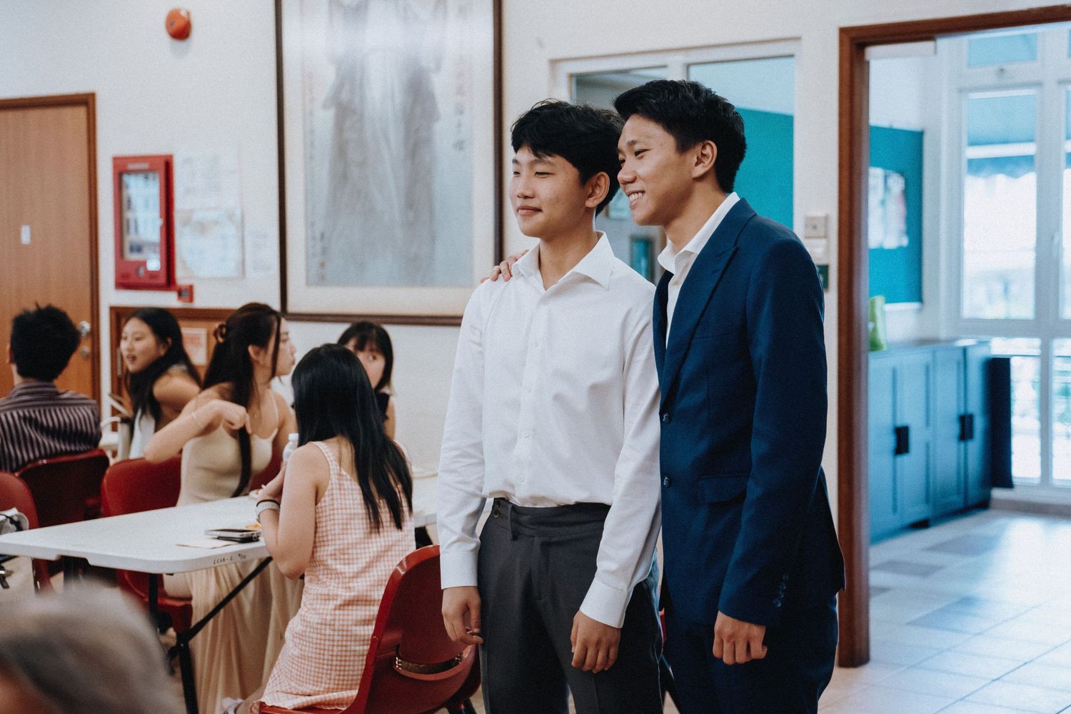 Two young men in formal attire stand together at a wedding reception venue.